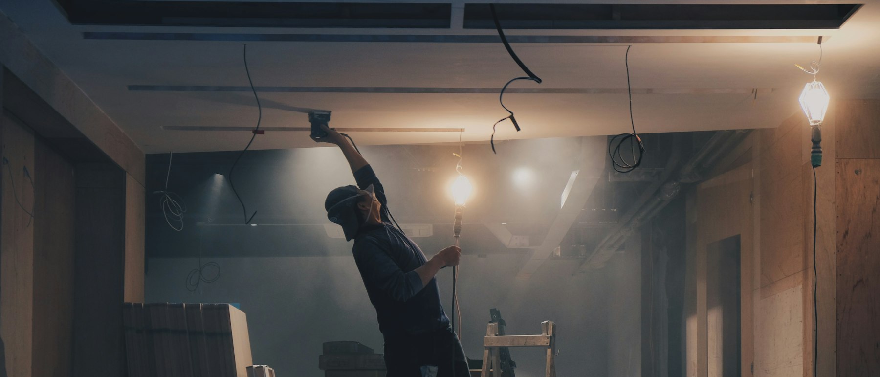 Worker on a ladder sanding a ceiling in an unfinished interior, dust caught in the work lights